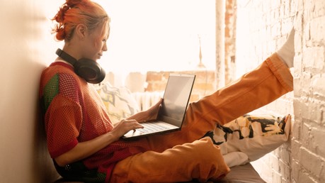 A young woman with orange hair sits comfortably near a window with over-the-ear headphones around her neck looking at her Windows 11 laptop.