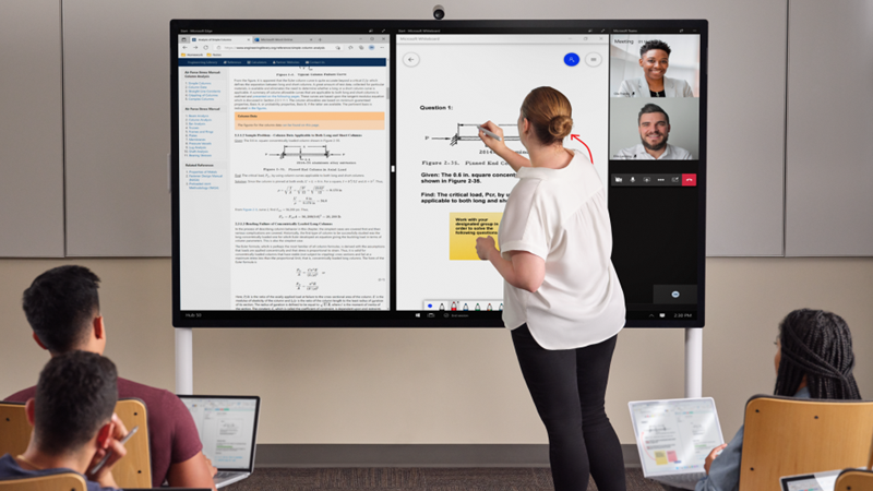 A teacher is at the front of a class inking on a Hub 85” with a light gray Surface Pen. Edge Whiteboard and Teams is on the screen. In the foreground several student have a Pro 7+ and are taking notes in One Note.